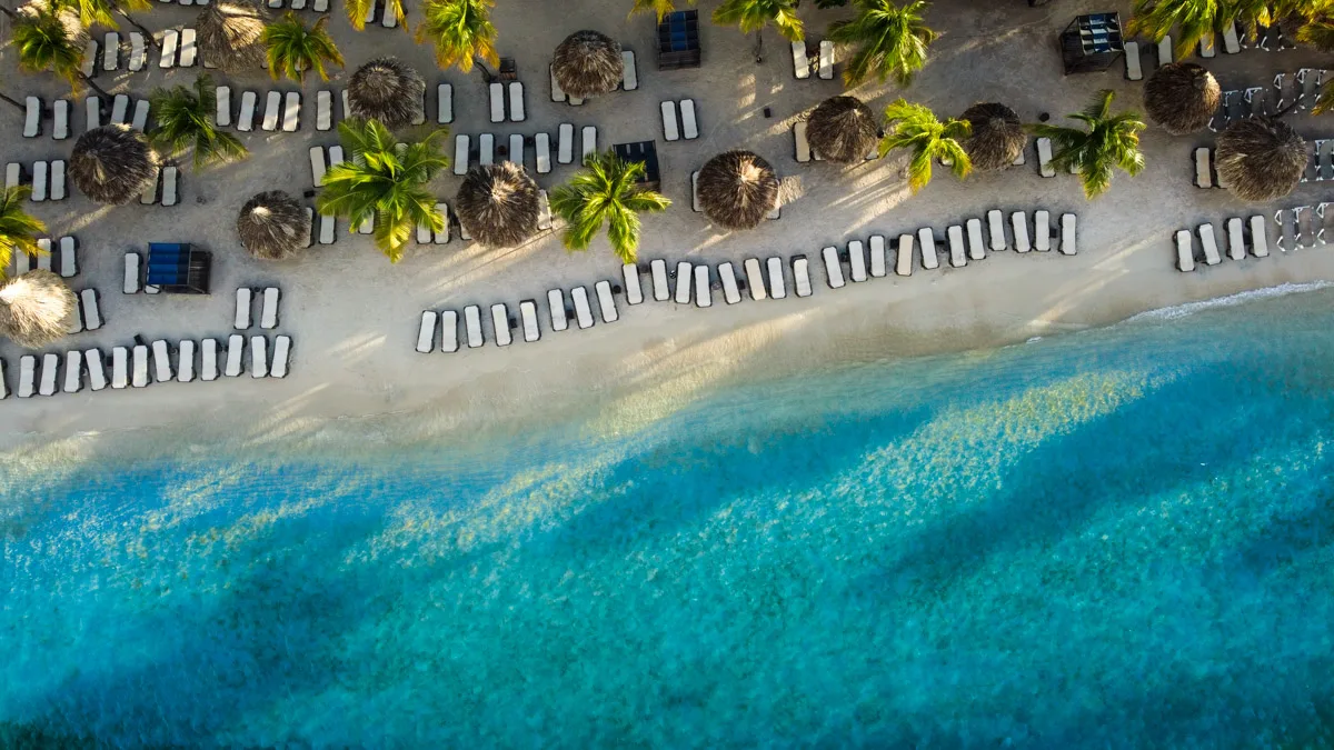 High-angle shot over the crystal-clear waters of Mambo Beach in Curaçao, highlighting the gradient of blues in the sea with beach umbrellas and palm trees adorning the shoreline.