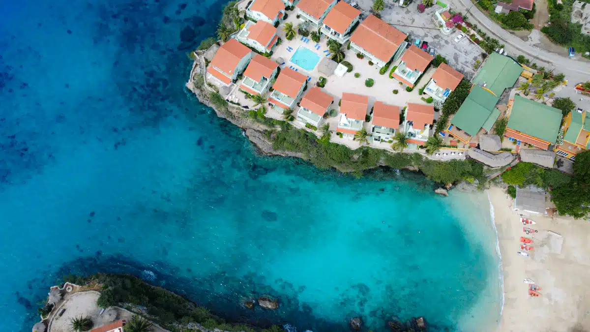 An aerial perspective of a picturesque Curacao coastal community, featuring cliffside houses with ocean views, nestled near lush greenery and clear blue waters, ideal for diving enthusiasts.