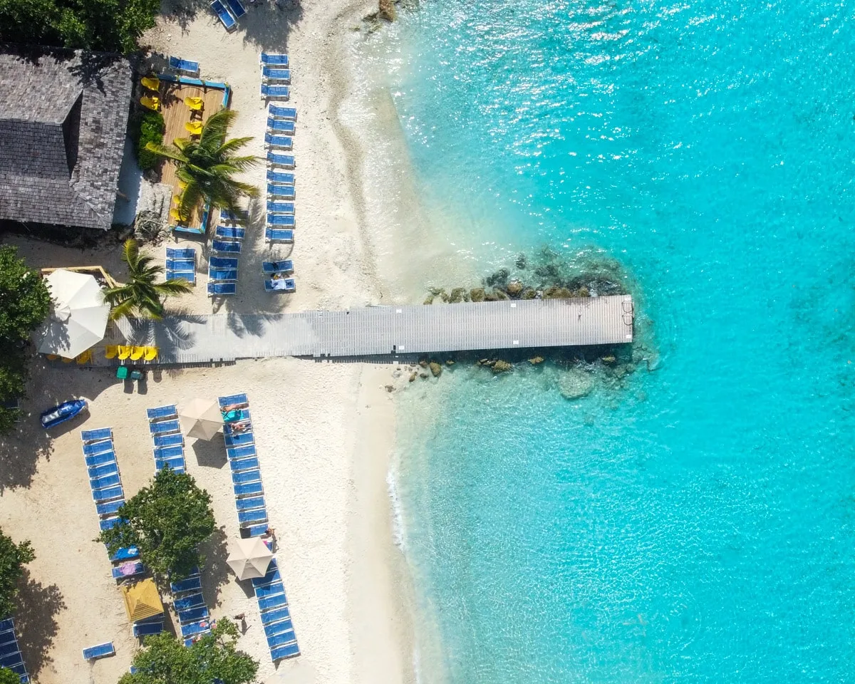 A wooden pier extends into the crystal-clear turquoise sea at a Curaçao beach resort, framed by rows of sun loungers and vibrant kayaks, inviting relaxation and water activities