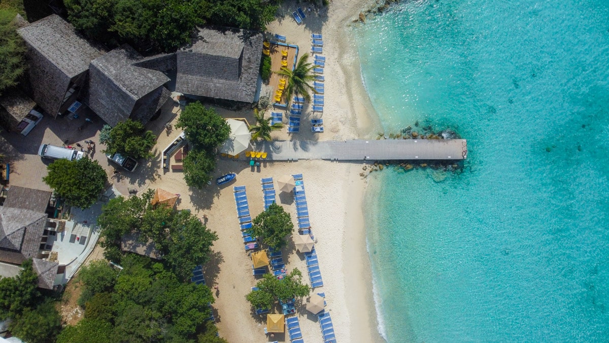 Bird's-eye view of a sandy beach with rows of blue lounge chairs under yellow umbrellas, next to a wooden pier extending into clear blue waters.