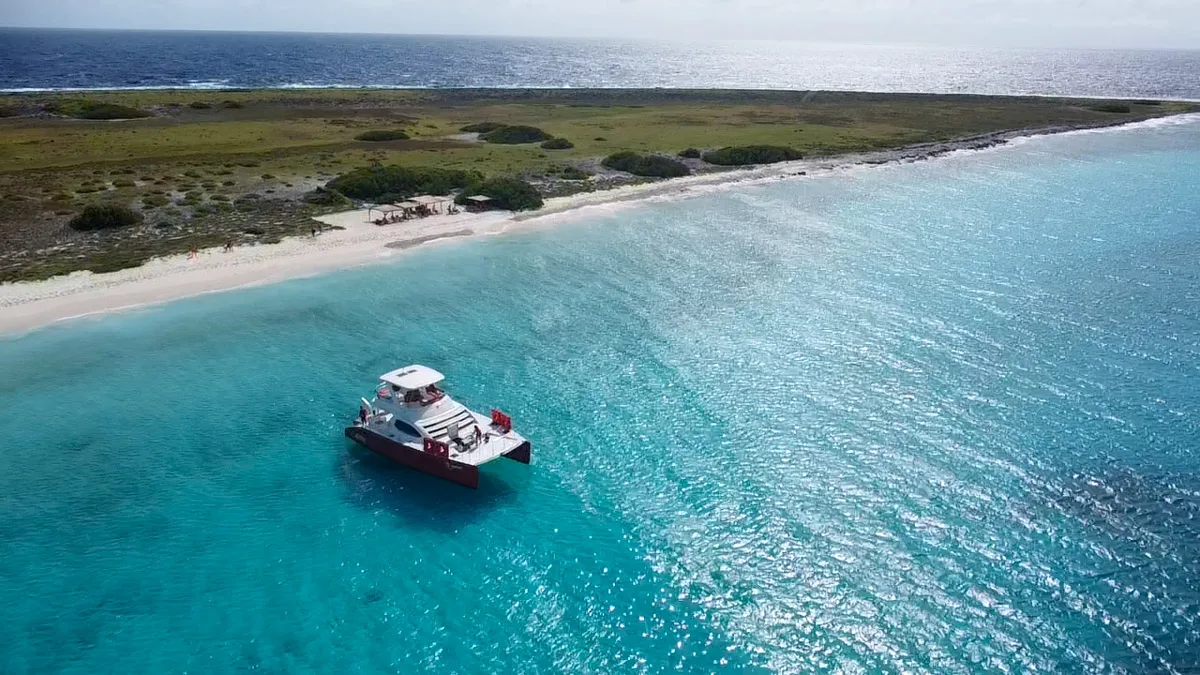 A bright red and white boat is moored off a secluded beach with crystal-clear blue waters, under a clear sky, perfect for Klein Curacao boat trips.