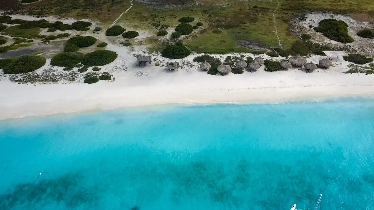 Aerial view of a stunning white sand beach bordering clear turquoise waters, with green shrubbery and a few small huts scattered along the shoreline.