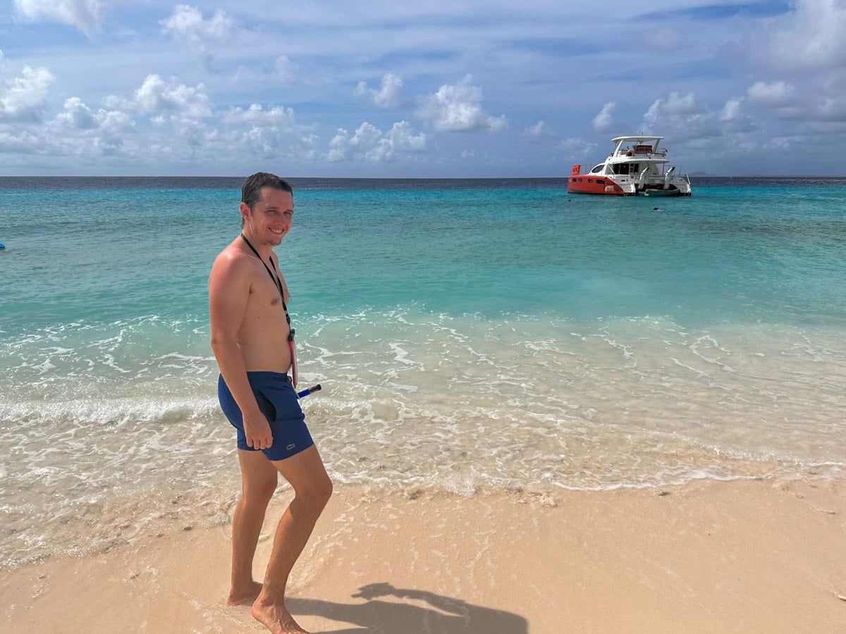 the authors husband smiling at the camera, standing on a white sandy beach with a red and white boat in the background, depicting a classic Klein Curacao boat trip scene.