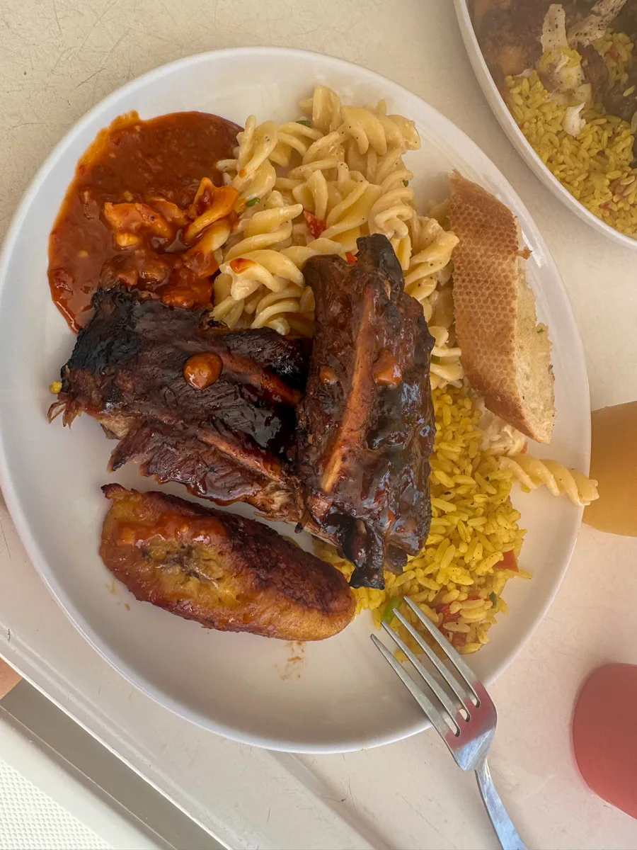 A plate with a hearty meal consisting of barbecued ribs, pasta, plantains, and a side of bread and rice, suggesting a fulfilling meal after a day of beach activities.