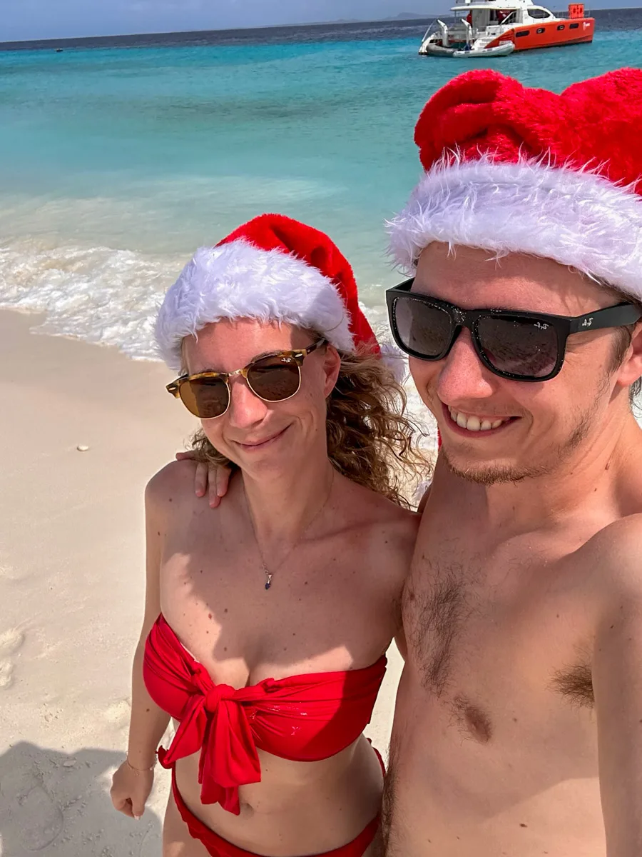 A couple in festive Santa hats poses for a selfie, with stunning clear blue waters and a red and white boat in the background, highlighting the joy of a Klein Curacao boat trip during the holiday season.