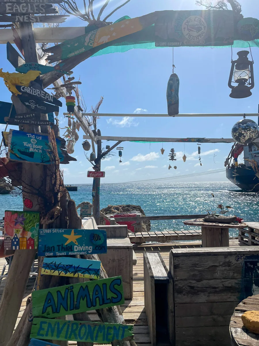 The rustic entrance to a beach bar in Curaçao, adorned with colorful signs and driftwood, framing a view of the shimmering Caribbean Sea, inviting the author and visitors to a unique beachside retreat