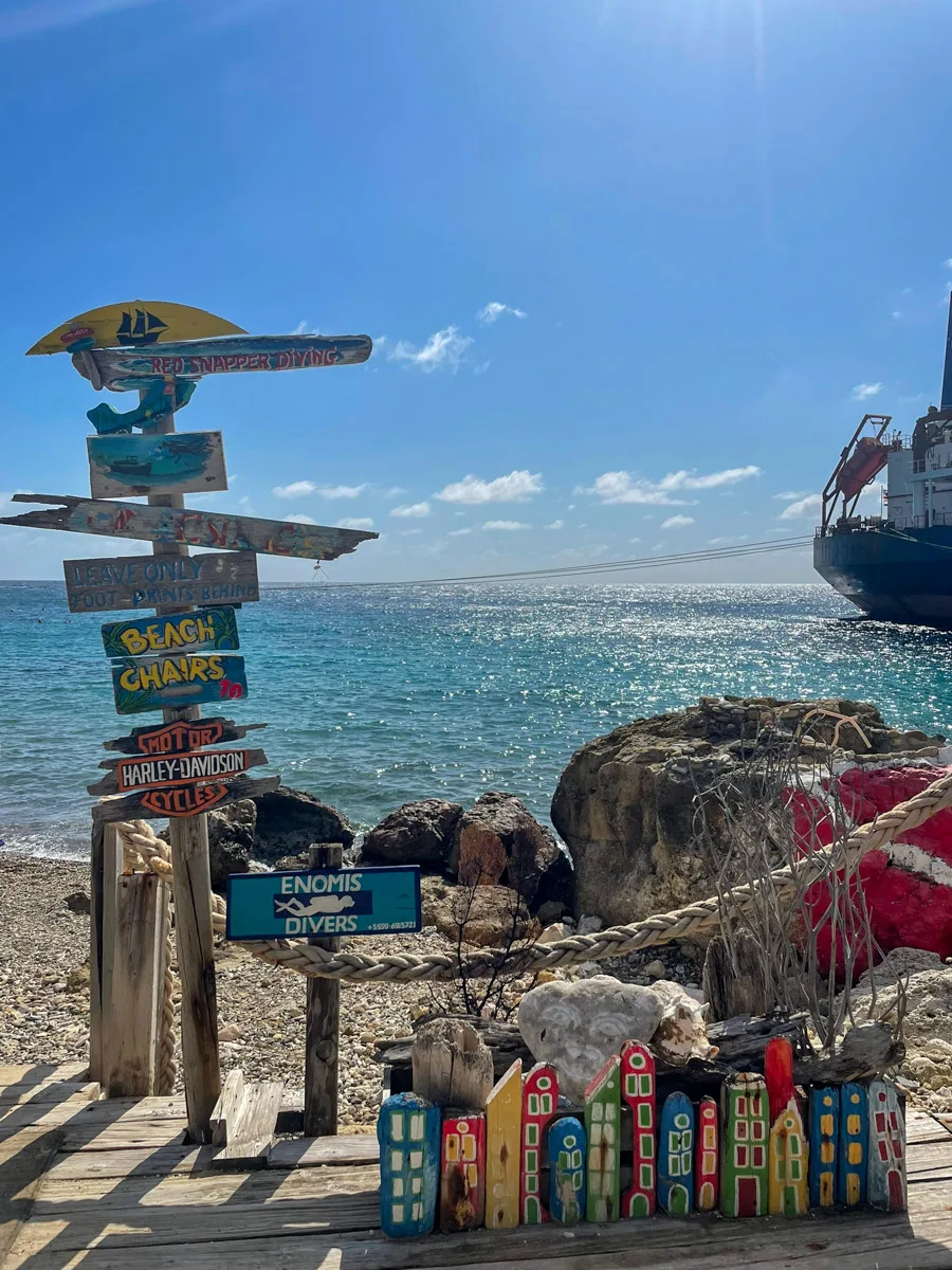 The rustic entrance to a beach bar in Curaçao, adorned with colorful signs and driftwood, framing a view of the shimmering Caribbean Sea, inviting the author and visitors to a unique beachside retreat
