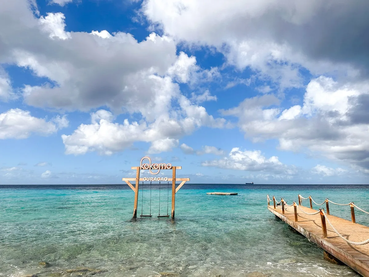 The iconic Kokomo Beach sign stands on a wooden dock over the clear blue waters of Curaçao, a popular spot for visitors to capture memories