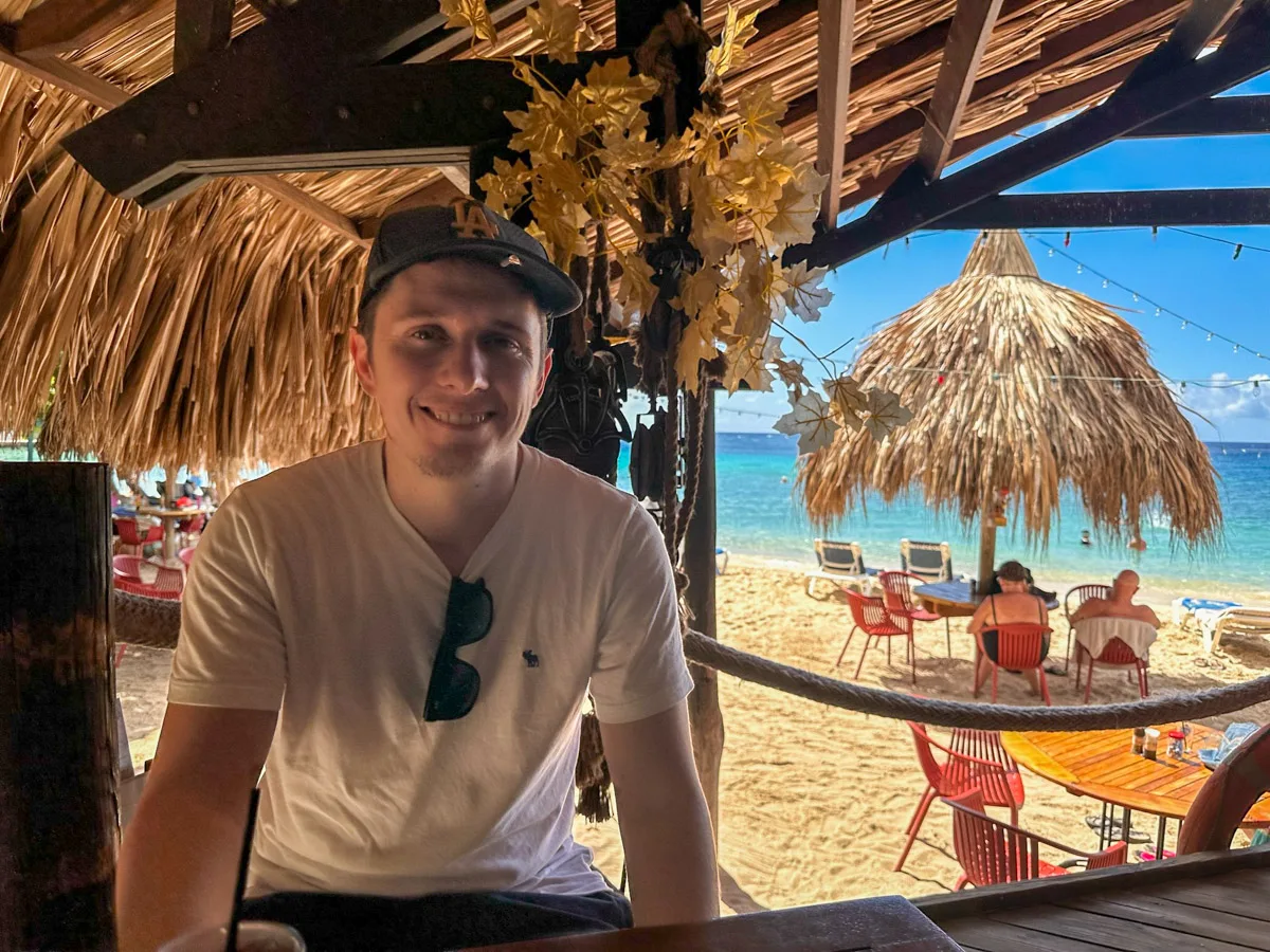 A smiling man sits at a beach bar in Curaçao, with a backdrop of a sandy beach and crystal-clear waters, reflecting the laid-back island lifestyle. you can see the authors husband in the picture sitting in piscaderabaai, one of the best things to do in curacao
