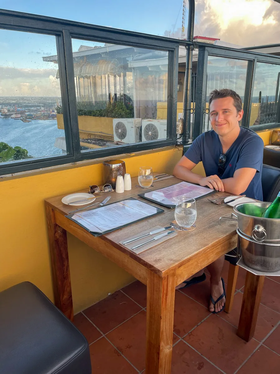 The author's husband awaits a meal at a rooftop restaurant in Curaçao, with a stunning view of the harbor and the island's vibrant cityscape at dusk.