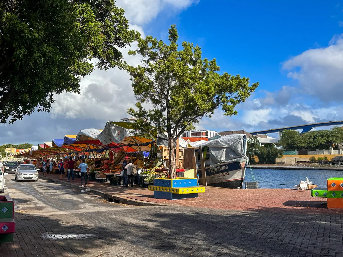 A bustling open-air market in Curaçao, lined with colorful produce stands under shaded canopies, with the floating market boats moored along the waterfront