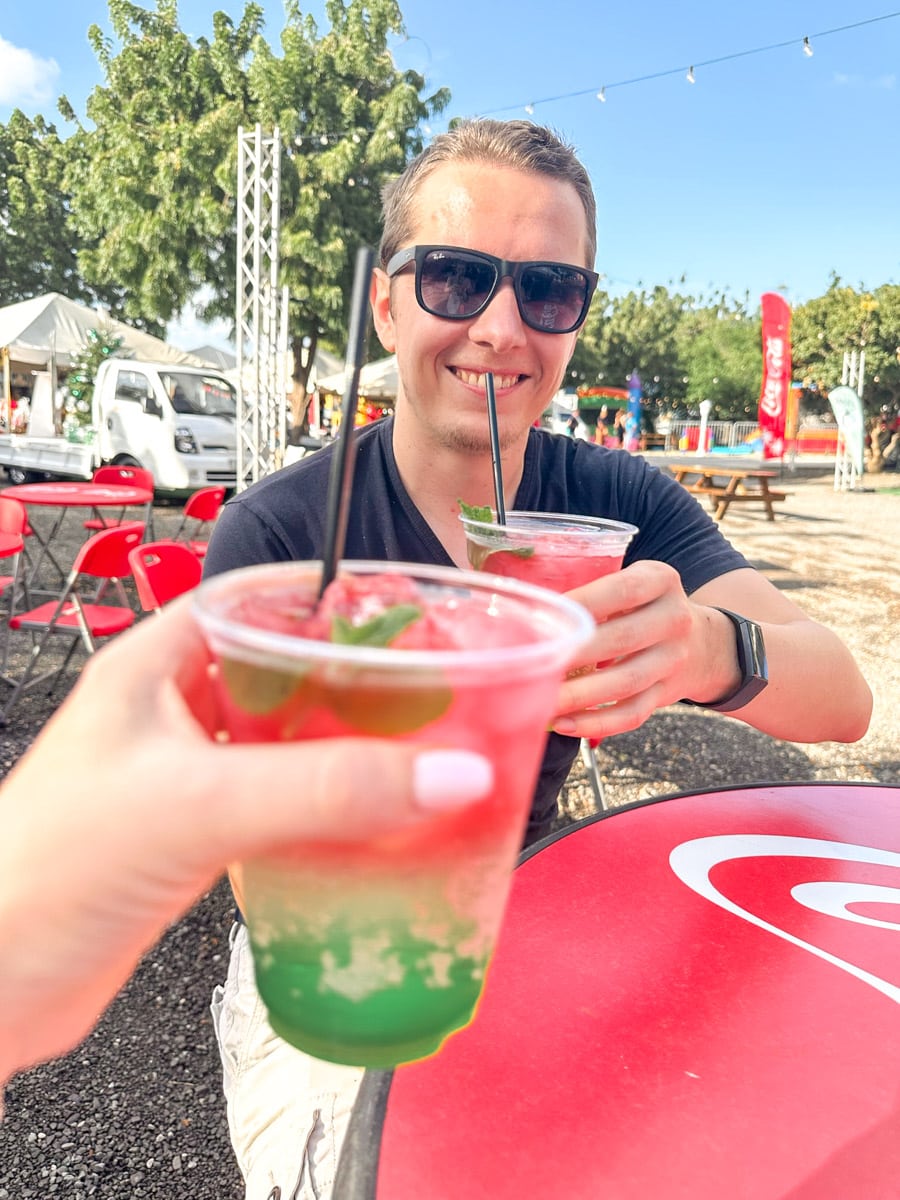 The authors husband enjoying a colorful layered cocktail in a casual outdoor setting, with a festive atmosphere and market stalls in the background.