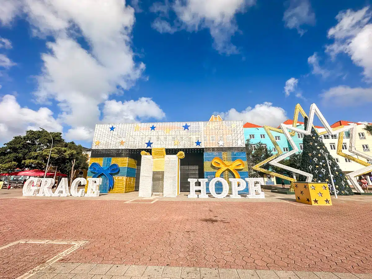 A large festive outdoor display spelling "GRACE HOPE" in big letters, with oversized gift box and star decorations under a sunny sky, in a square lined with colorful buildings.