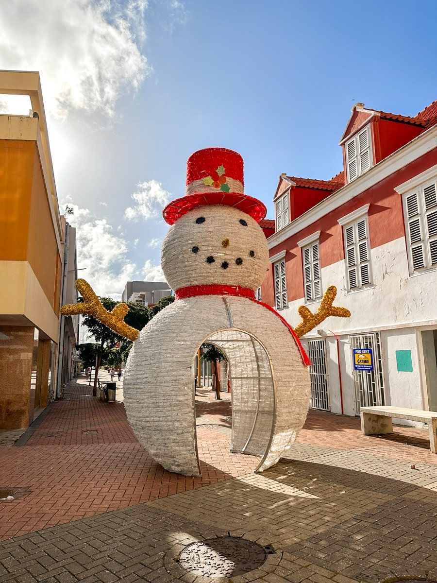 A giant snowman sculpture wearing a red hat and scarf adorns a city street, bringing a touch of whimsy to the tropical setting. totally lovely Christmas decoration in Curacao in December