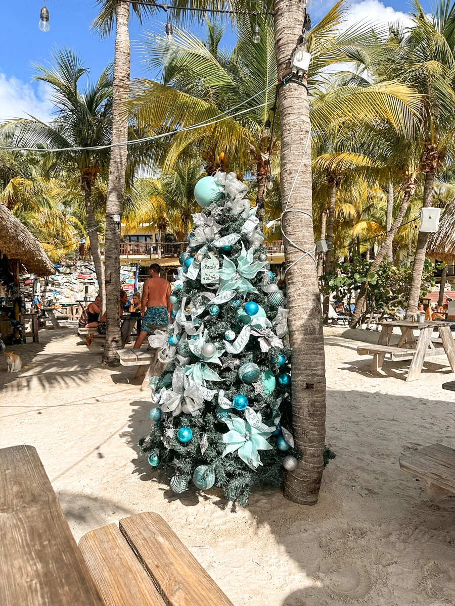 A Christmas tree decorated with blue and silver ornaments stands on a sandy beach, surrounded by palm trees and beachgoers.