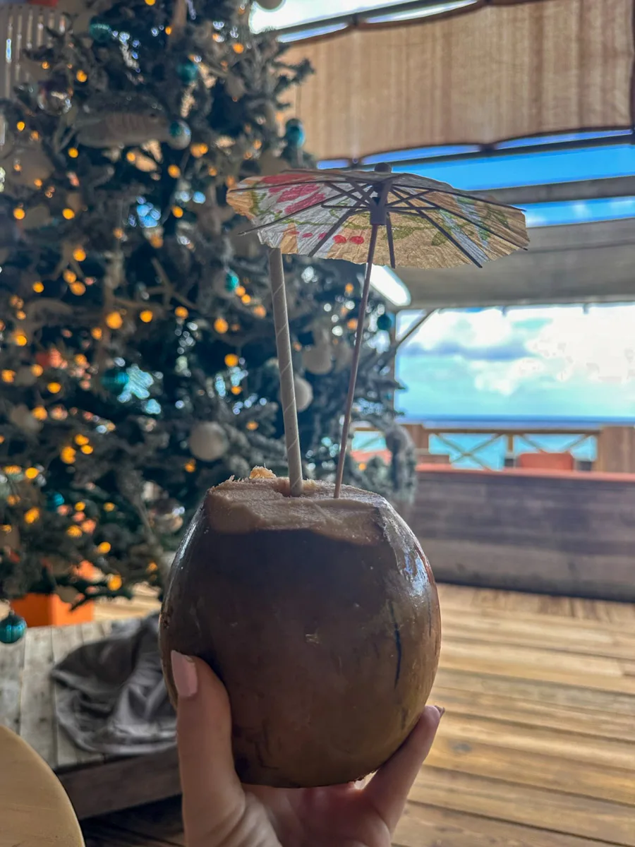 A festive drink served in a coconut shell with a mini umbrella, placed on a wooden table beside a small Christmas tree, with a blurred ocean view in the background. Christmas at Kokomo Beach Curacao