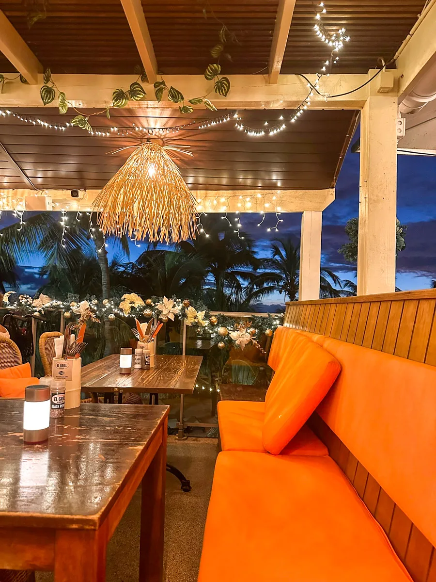 An outdoor seating area at dusk, featuring orange cushions, twinkling fairy lights, and a tropical ambiance with a view of the ocean at El Grill in Curacao for Christmas