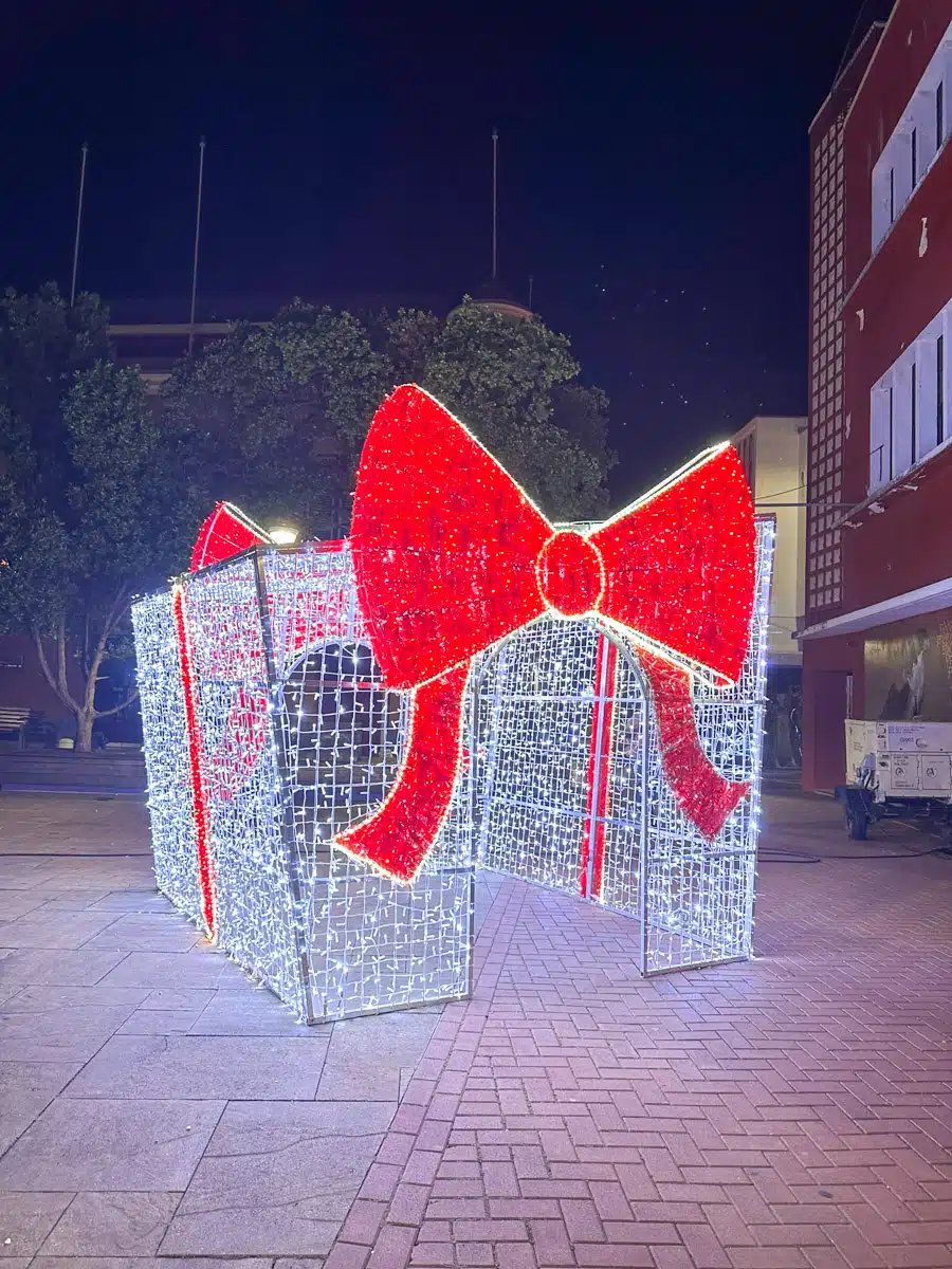 A large, glowing red Christmas bow decoration on a public square at night, adding a festive touch to the urban setting.