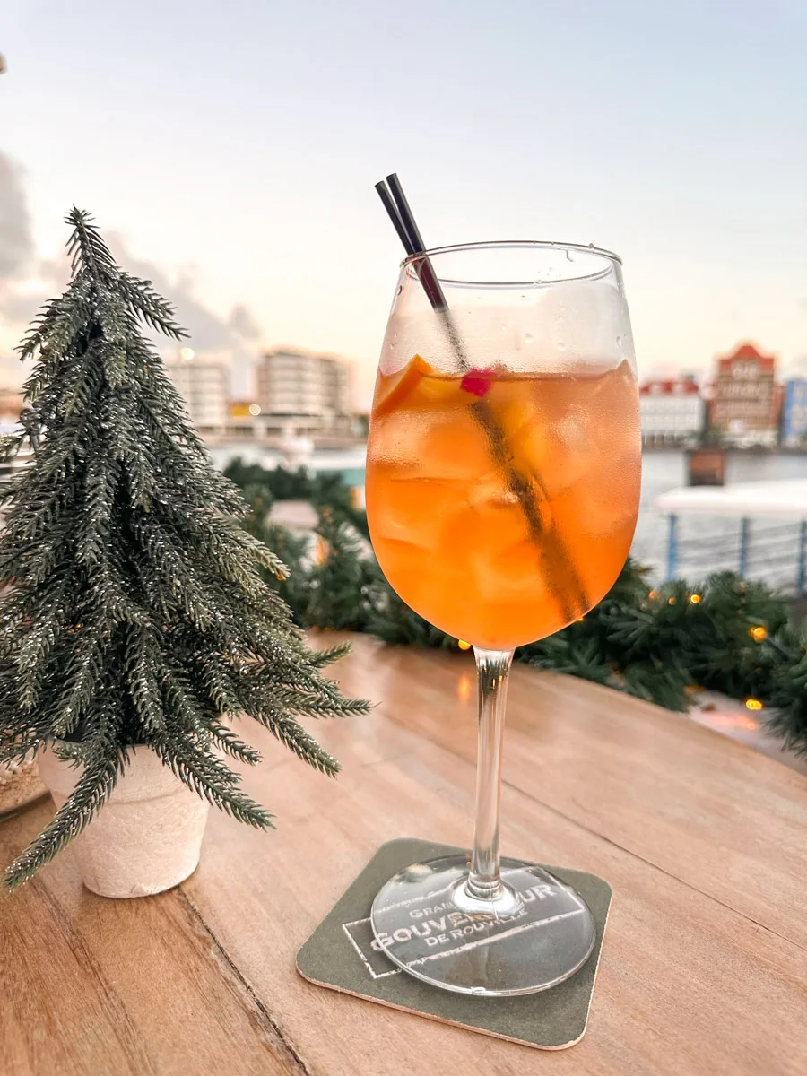 A refreshing cocktail with an orange hue, served in a wine glass and garnished with a slice of orange, placed on a wooden table with a mini Christmas tree.