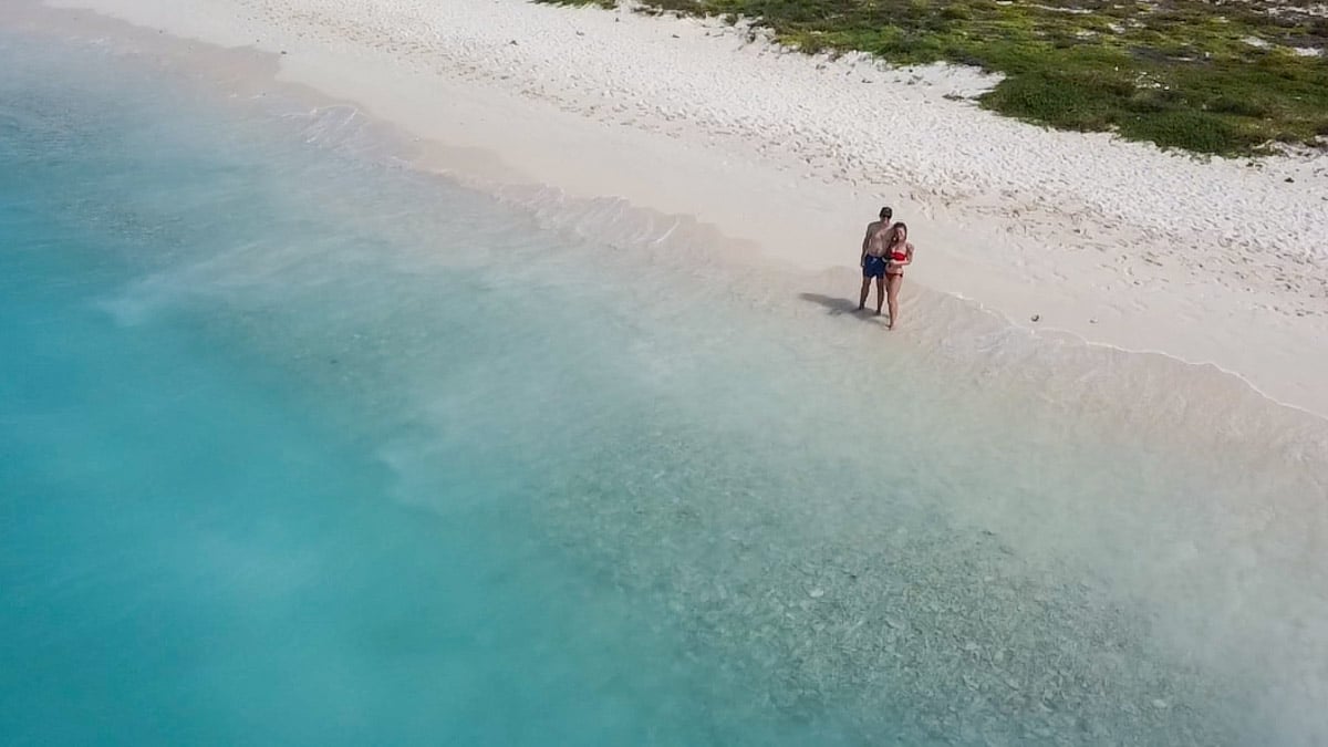 A couple stands close together on a pristine white sand beach, with crystal-clear turquoise waters gently lapping at their feet, under a bright tropical sun.