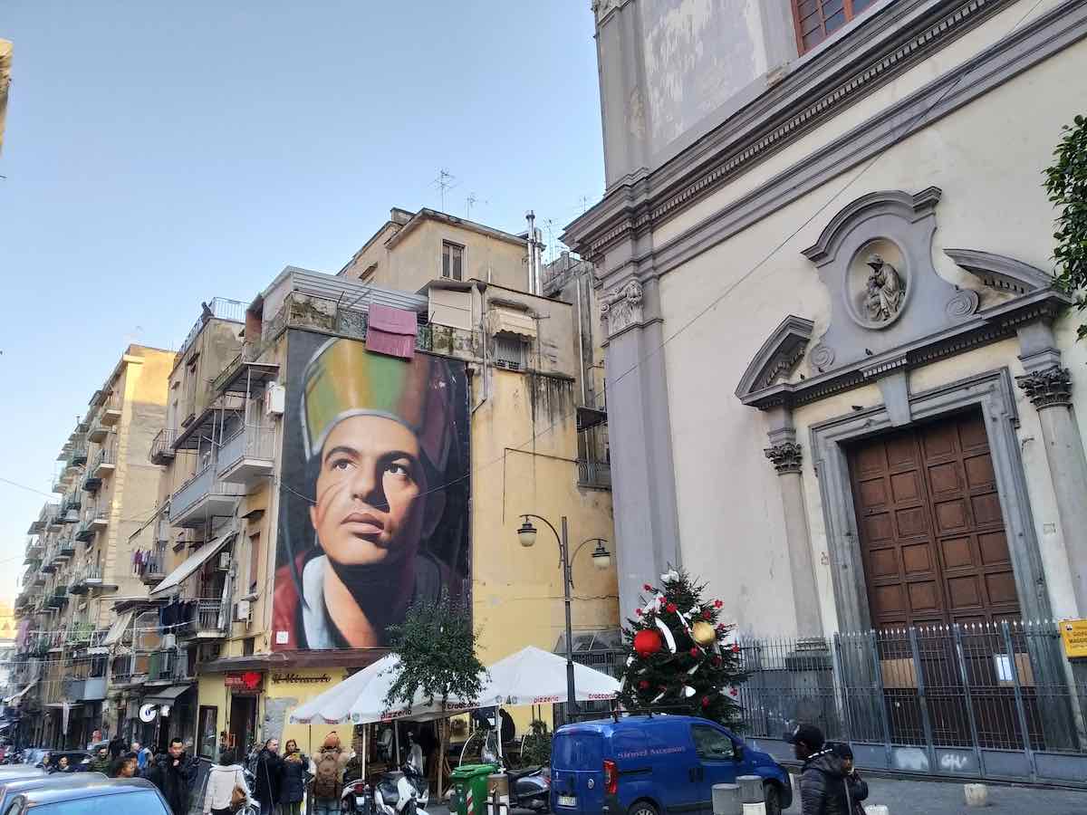 A large mural of Saint Gennaro, the patron saint of Naples, painted on the side of a building, overlooking a street scene with pedestrians and a church to the right.