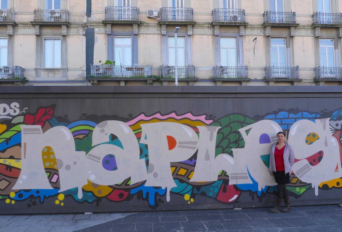 A traveler stands before a vibrant street mural spelling out 'Naples' with colorful and artistic flair, capturing the city's lively urban culture.