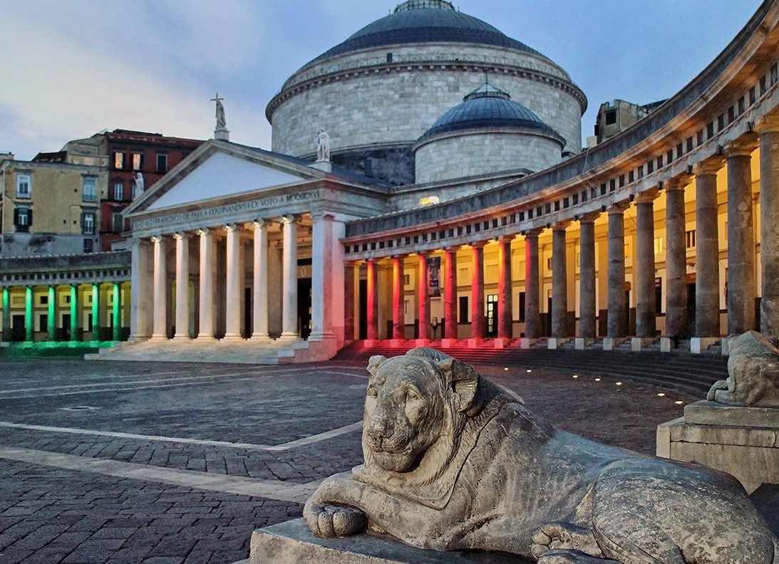 Twilight descends on Piazza del Plebiscito in Naples, with the colonnaded facade of the Basilica Royal lit in the tricolor, and ancient stone lions standing guard