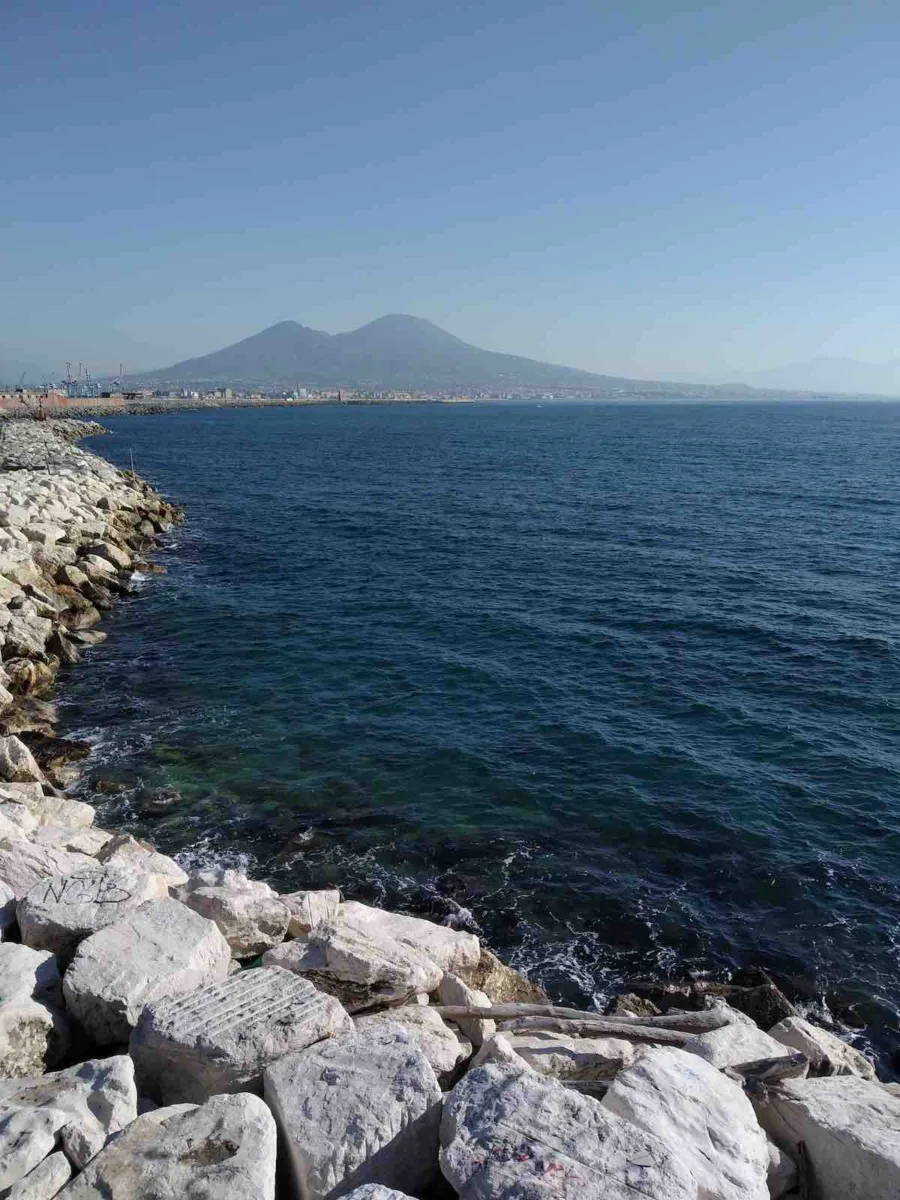 "A serene view of Mount Vesuvius across the bay from Naples, with the calm blue waters in the foreground and the city’s silhouette under the mountain’s watchful presence."
