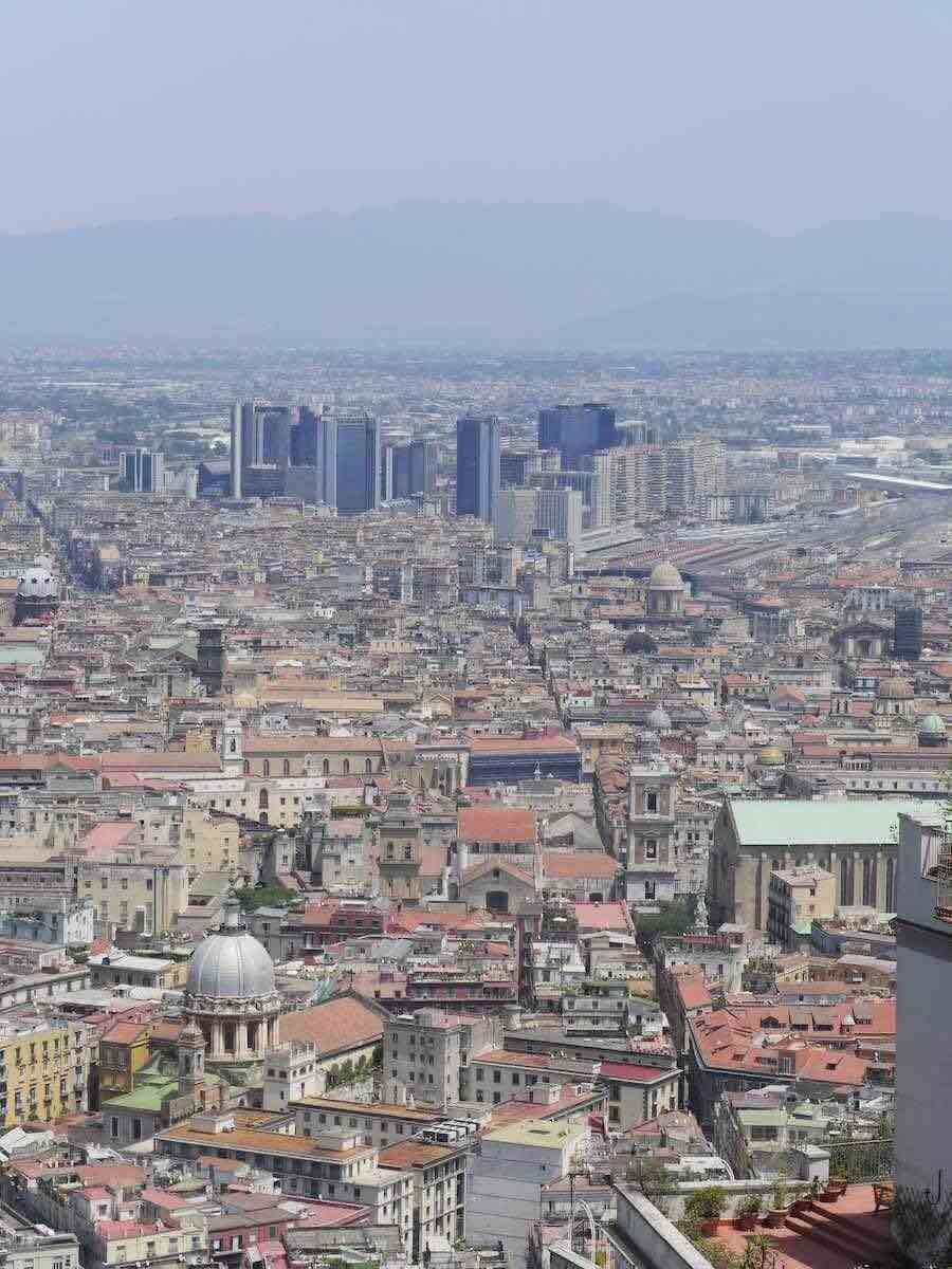 An expansive aerial view of Naples showing the dense urban landscape, with the prominent line of the Spaccanapoli street cutting through the city’s historical center