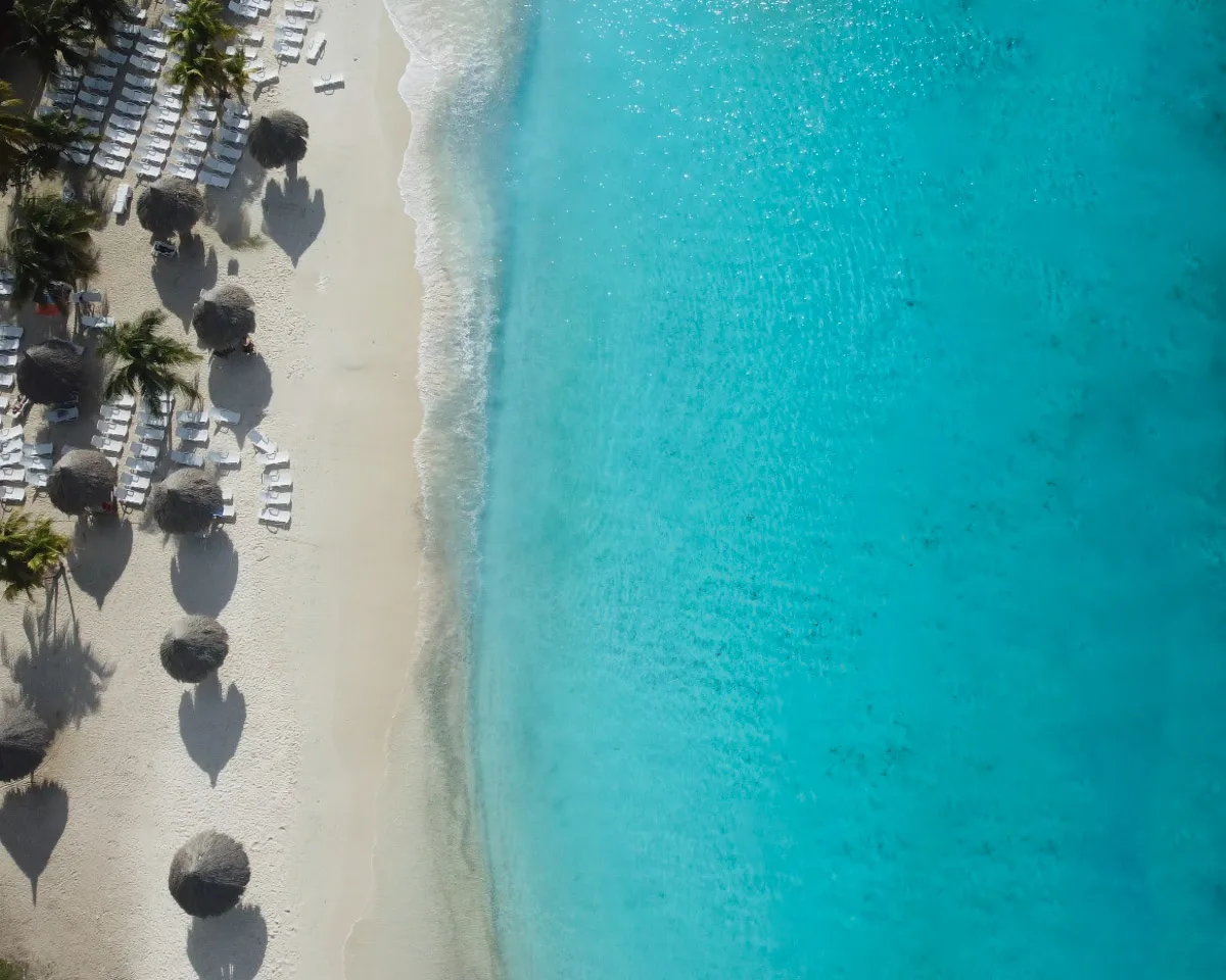 A closer aerial perspective of the beach highlighting the clear blue sea with patterns of waves near the shore, sun loungers neatly arranged under thatched umbrellas on the pristine sand.