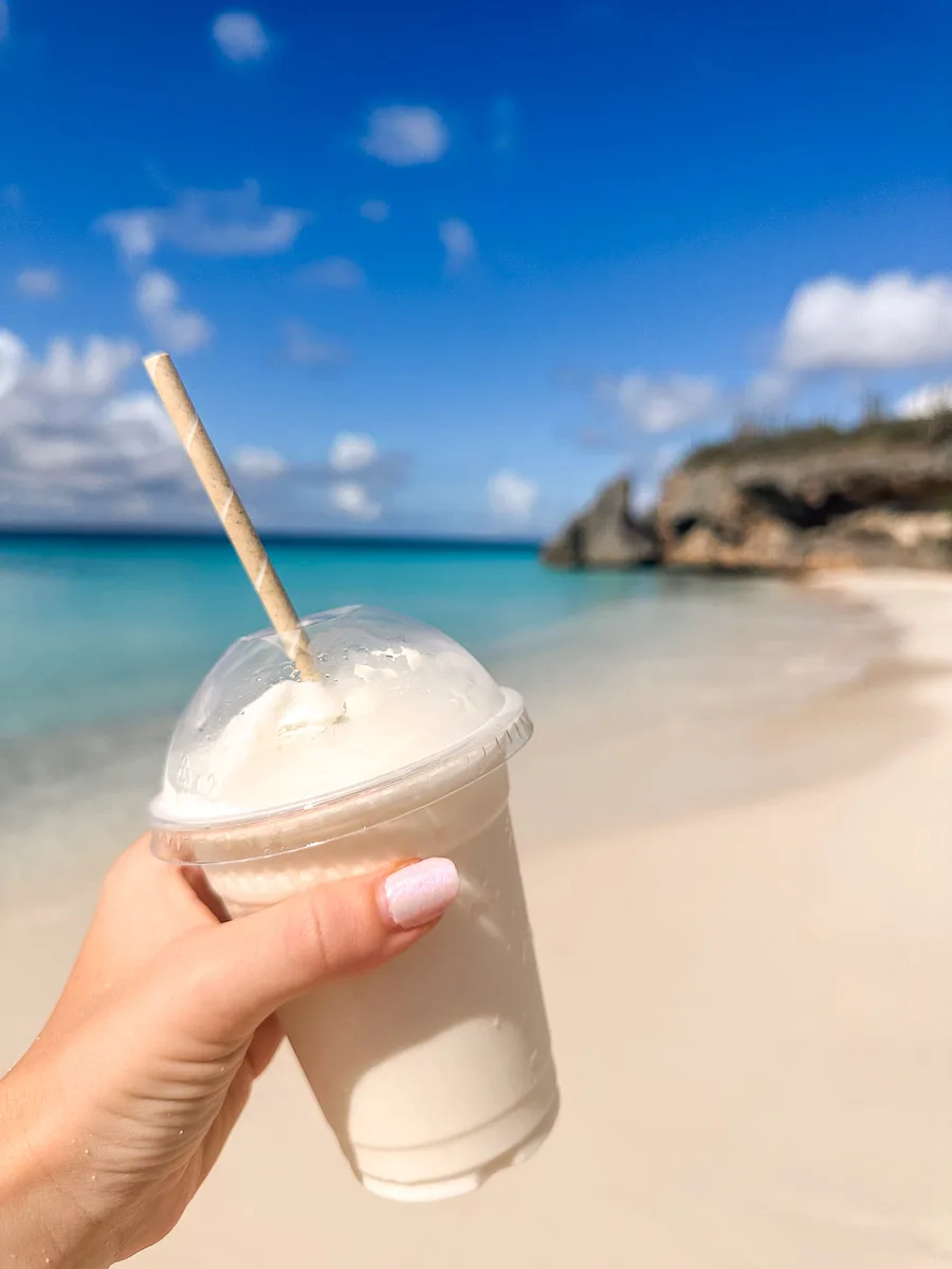 A hand holding a frothy iced drink with a biodegradable straw, with the backdrop of a serene blue sea and a sunny sky, suggesting a refreshing moment on a tropical beach.
