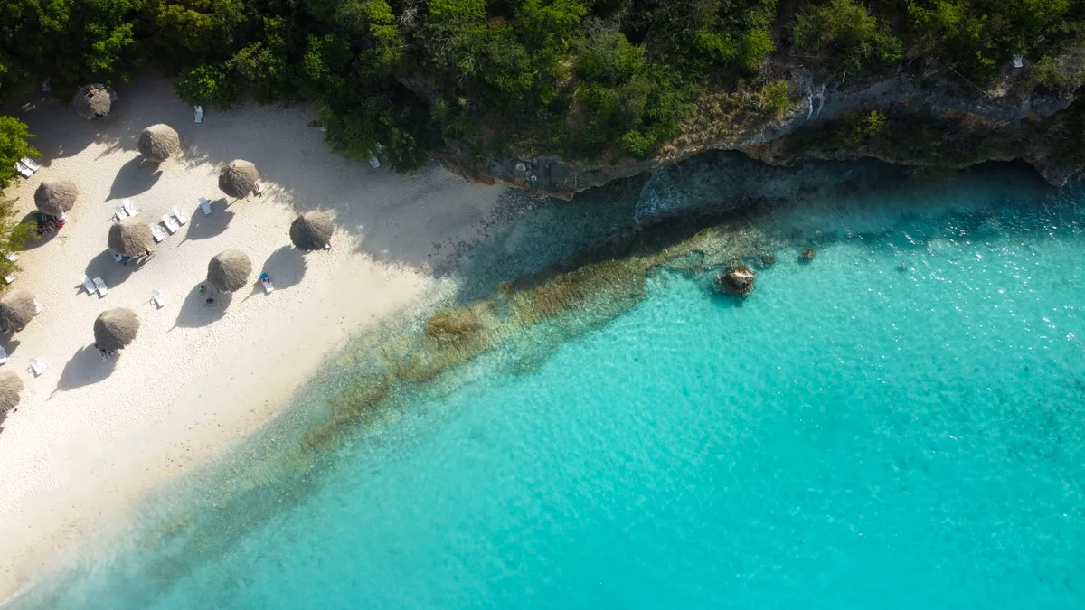 A high-angle drone shot of Cas Abao Beach showcasing the rugged coastline, clear turquoise waters, and a secluded sandy beach with umbrellas, surrounded by lush greenery.