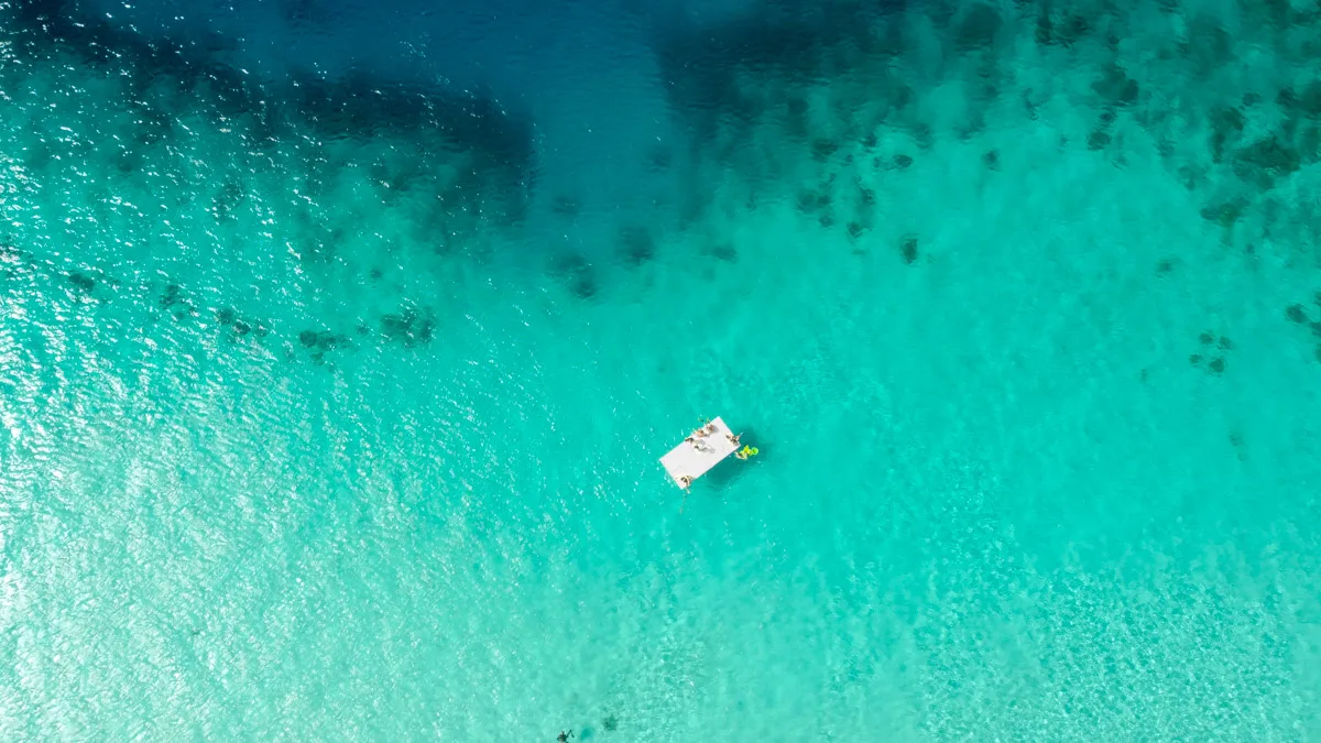 Overhead view of a boat floating on transparent turquoise waters near a white sandy beach with scattered sun loungers and parasols.