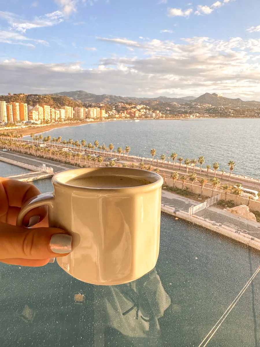 Close-up of a hand holding a coffee mug with a view of Malaga's coastline and palm tree-lined promenade reflected in the glass table.