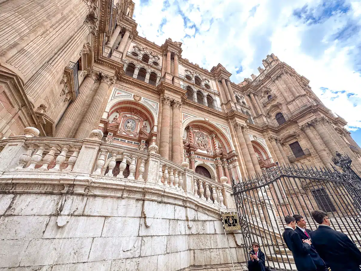 A close-up of Málaga Cathedral's ornate exterior with intricate architectural details and statues, under a blue sky with light clouds.