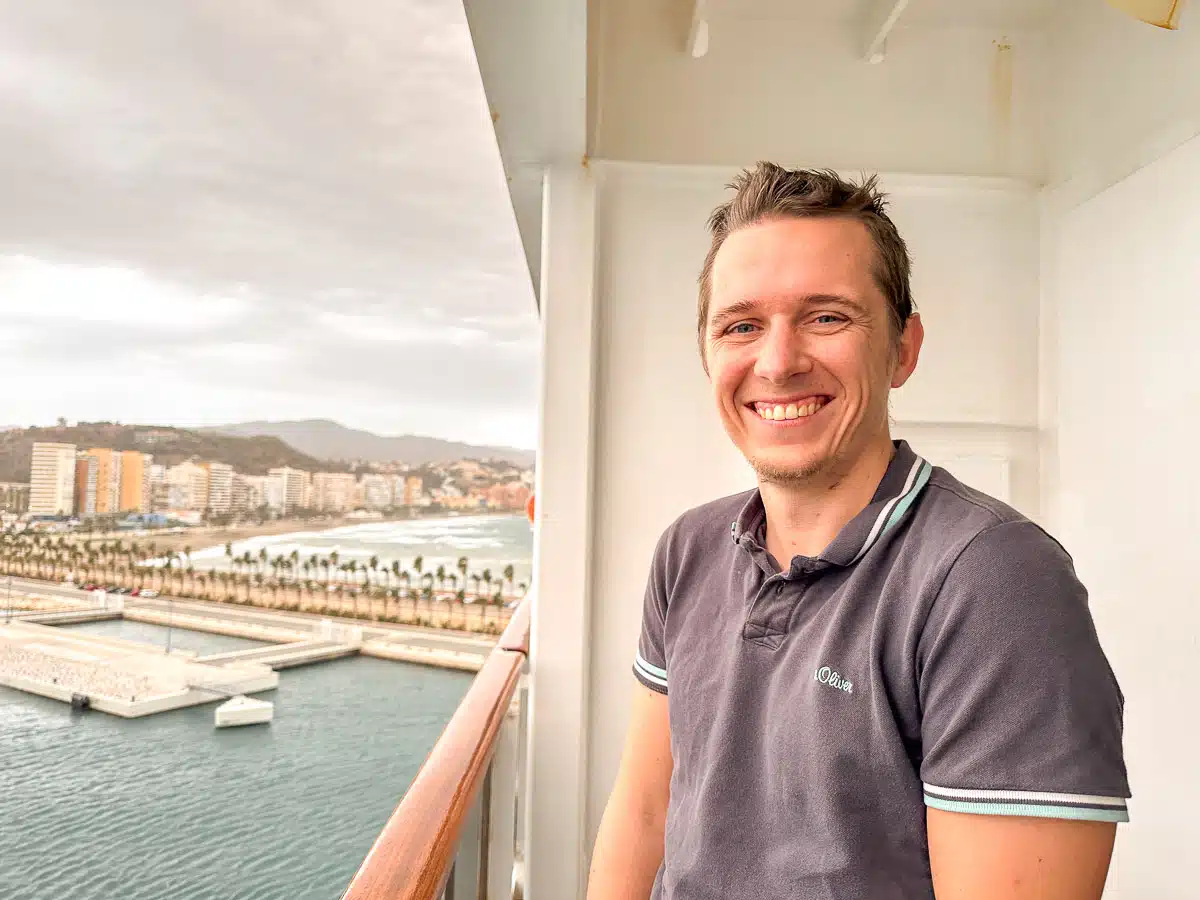 A smiling man standing on a cruise ship balcony overlooking Malaga's beachfront and port area with palm trees and mountains in the background
