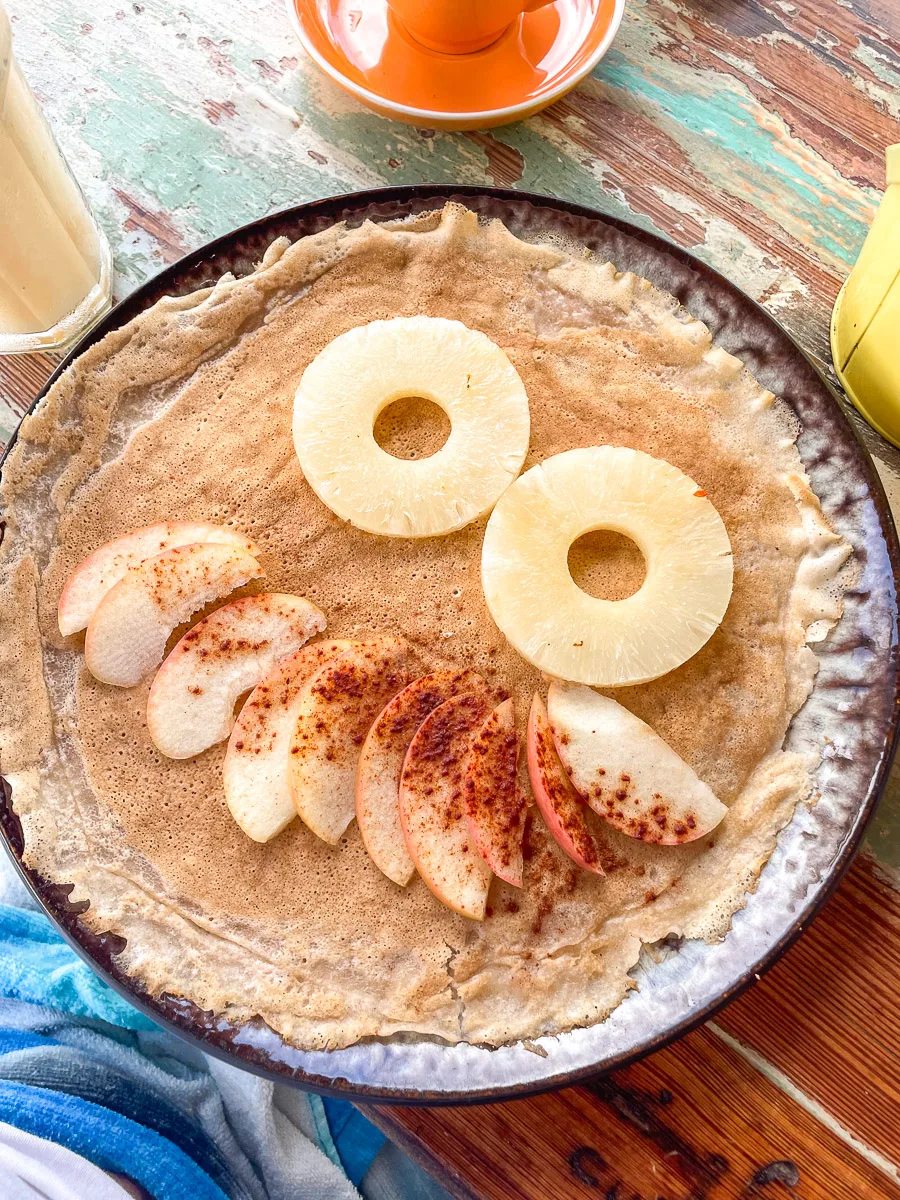 Close-up of a crepe with slices of pineapple and apple, sprinkled with cinnamon, on a rustic table with a vibrant orange cup and a refreshing drink in the background.