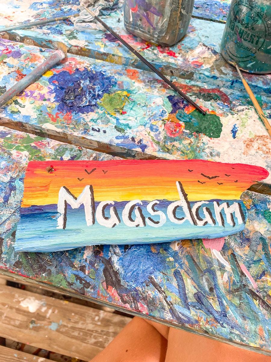 A colorful wooden sign with "Maasdam" painted in rainbow hues on a splattered and vibrant artist's palette, capturing the essence of creativity and fun.