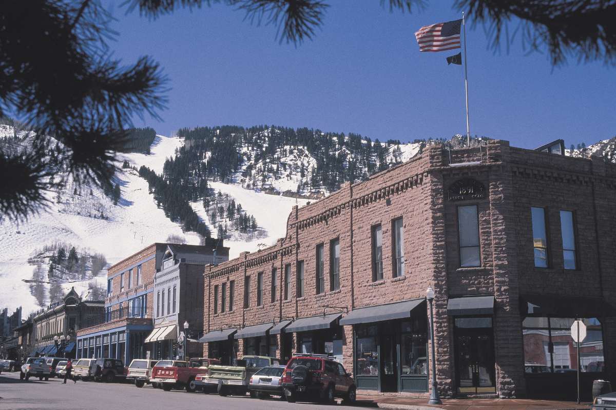A sunny street view in Aspen with charming brick buildings and the American flag fluttering, with snow-covered slopes in the distance. The cute inner city of Aspen makes it definitely worth visiting.