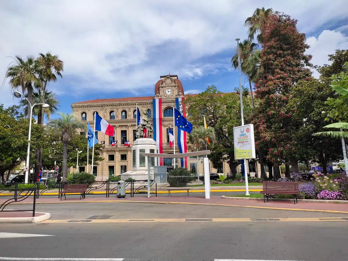 A daytime view of Cannes' city hall, an elegant building with French and European Union flags, in a square surrounded by lush greenery and vibrant flower beds.