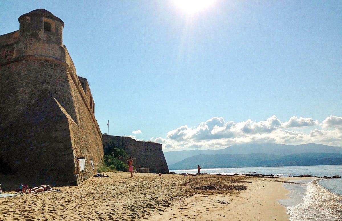 A sandy beach with an ancient round stone tower on the left, under a bright sun with a clear blue sky and mountains in the distance.