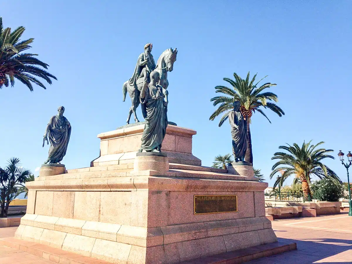 A grand monument featuring a bronze statue of a mounted figure flanked by statues and palm trees, under a clear blue sky. Beautiful Ajaccio monument you should definitely visit when one day in corsica