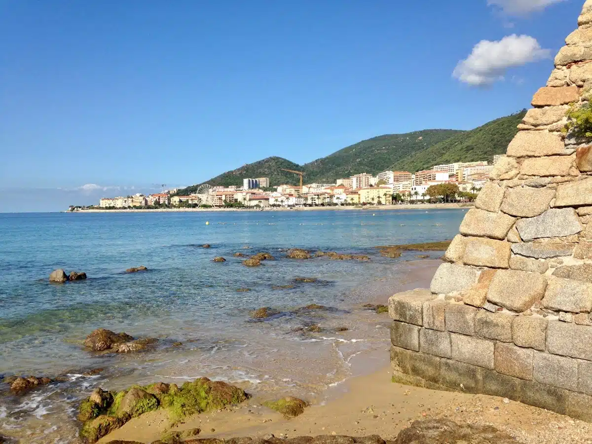 A coastal view showing a stone ruin in the foreground, with a calm sea, rocky shore, and a town in the background, all under a blue sky. Stunning beaches in Corsica should definitely be on your visits itinerary