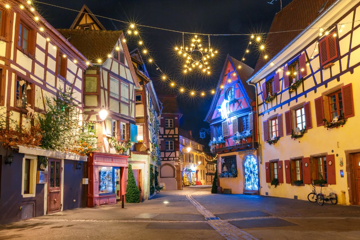 A festive evening scene in an Alsace town, with traditional half-timbered houses adorned with Christmas decorations and warm lights strung above the street, creating a cozy holiday atmosphere. One of the biggest reasons why Alsace is worth visiting