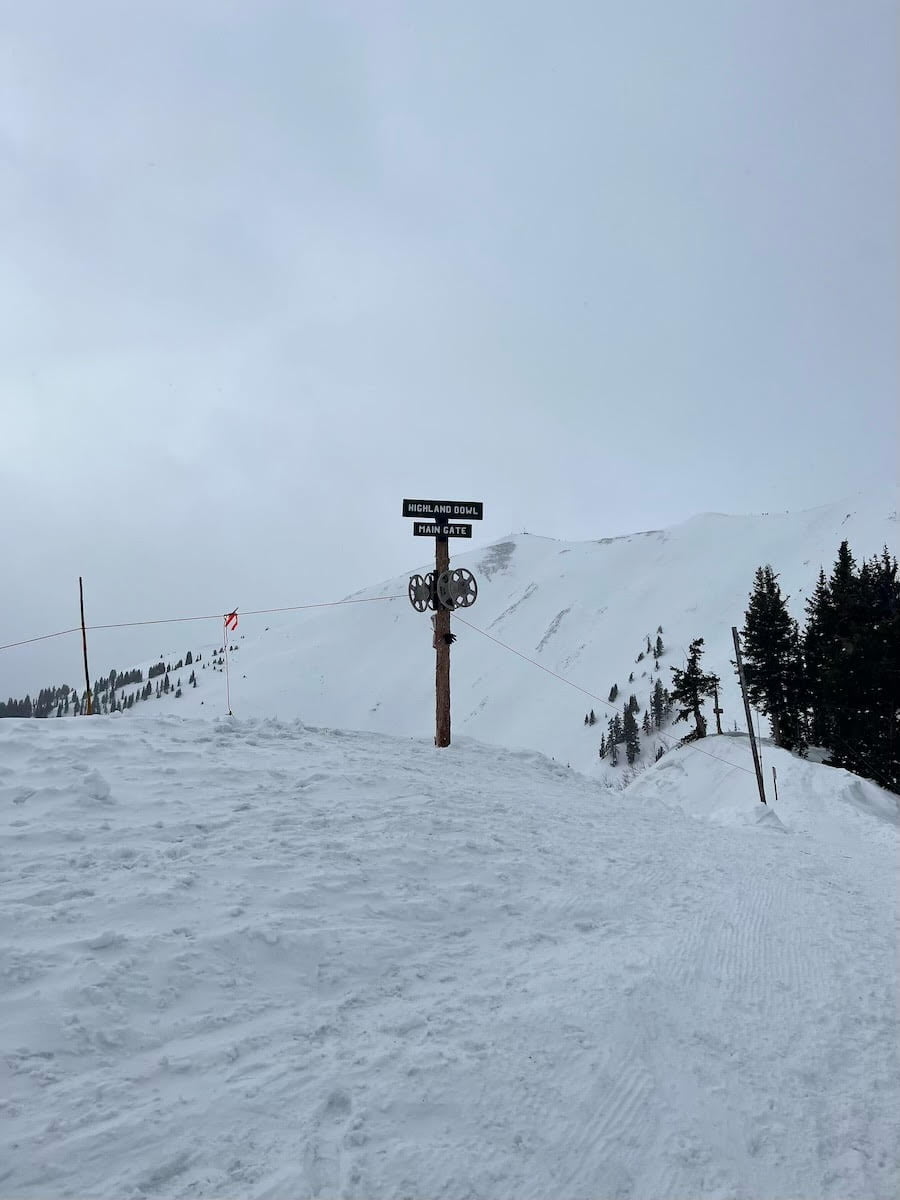 A signpost at the top of a snowy Aspen mountain with the text "Highland Bowl" indicating ski routes under an overcast sky.