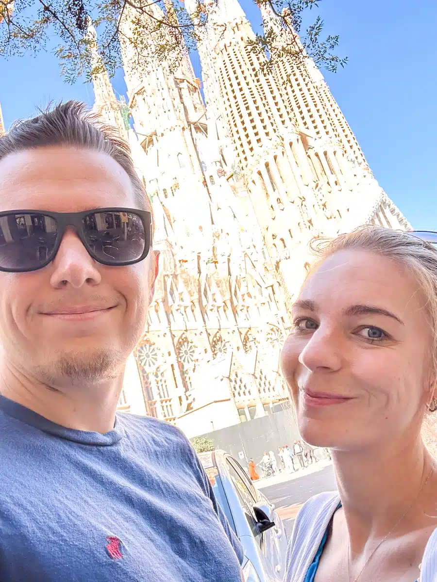 "A smiling couple taking a selfie with the towering spires of Barcelona's Sagrada Familia in the background under a clear blue sky. It is the author and her husband enjoying their one day in Barcelona from the cruise ship.