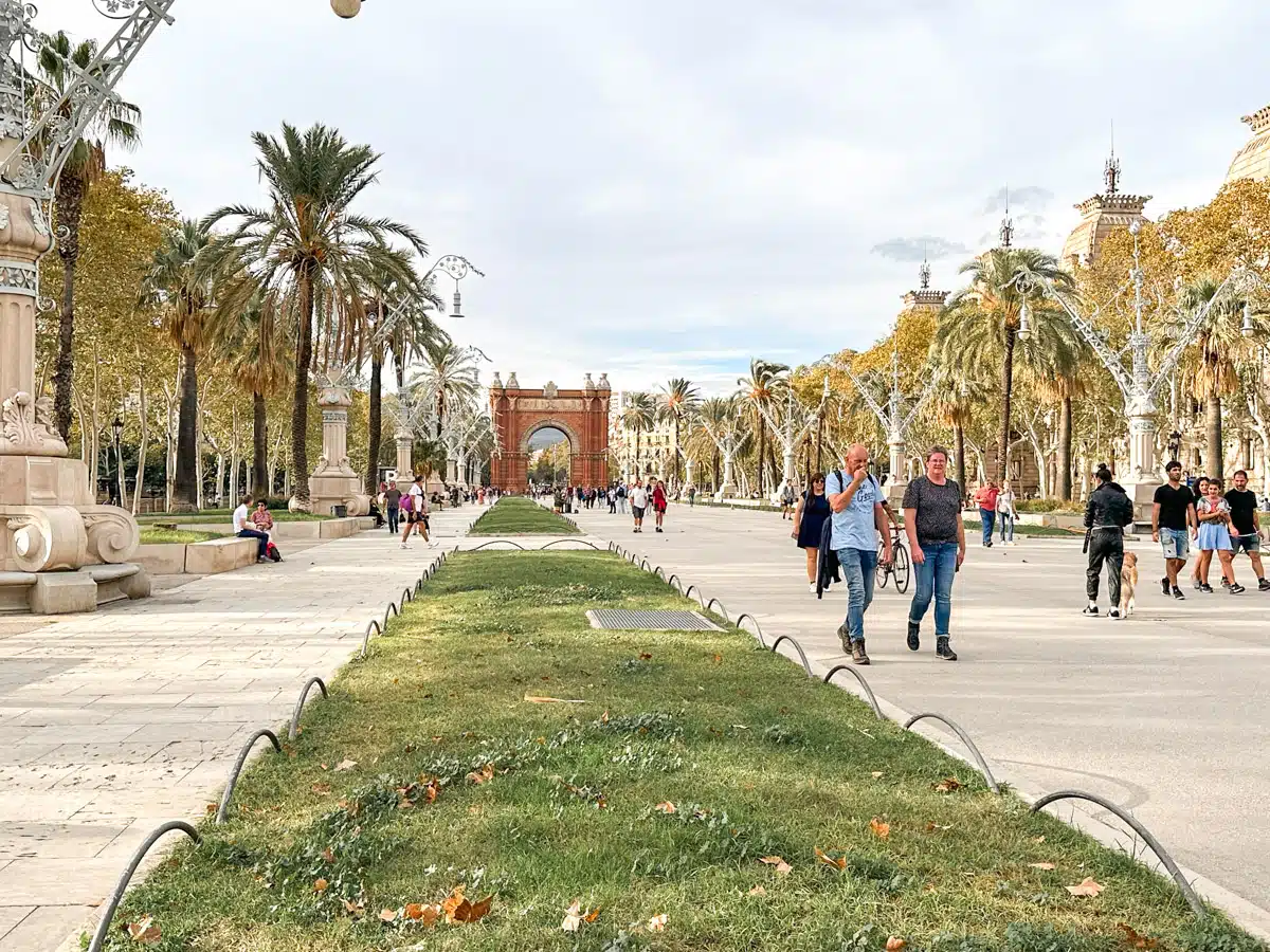 Visitors walking and relaxing along a wide tree-lined promenade in Parc de la Ciutadella, Barcelona, with the Triumphal Arch visible in the distance against a backdrop of clear blue sky.