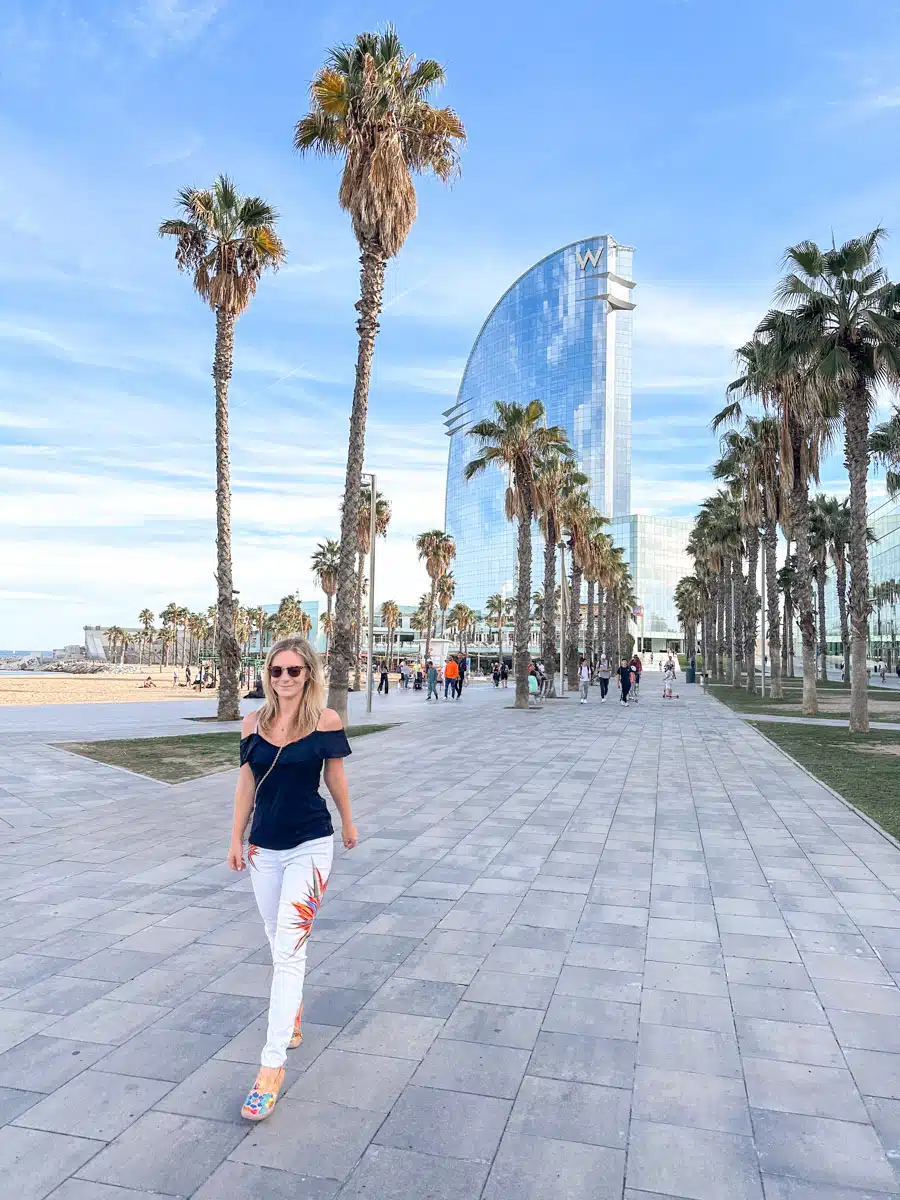 the author smiling as she strolls along the beachfront promenade in Barcelona, with the distinctive, modern silhouette of the W Hotel in the background and palm trees framing the clear blue sky.