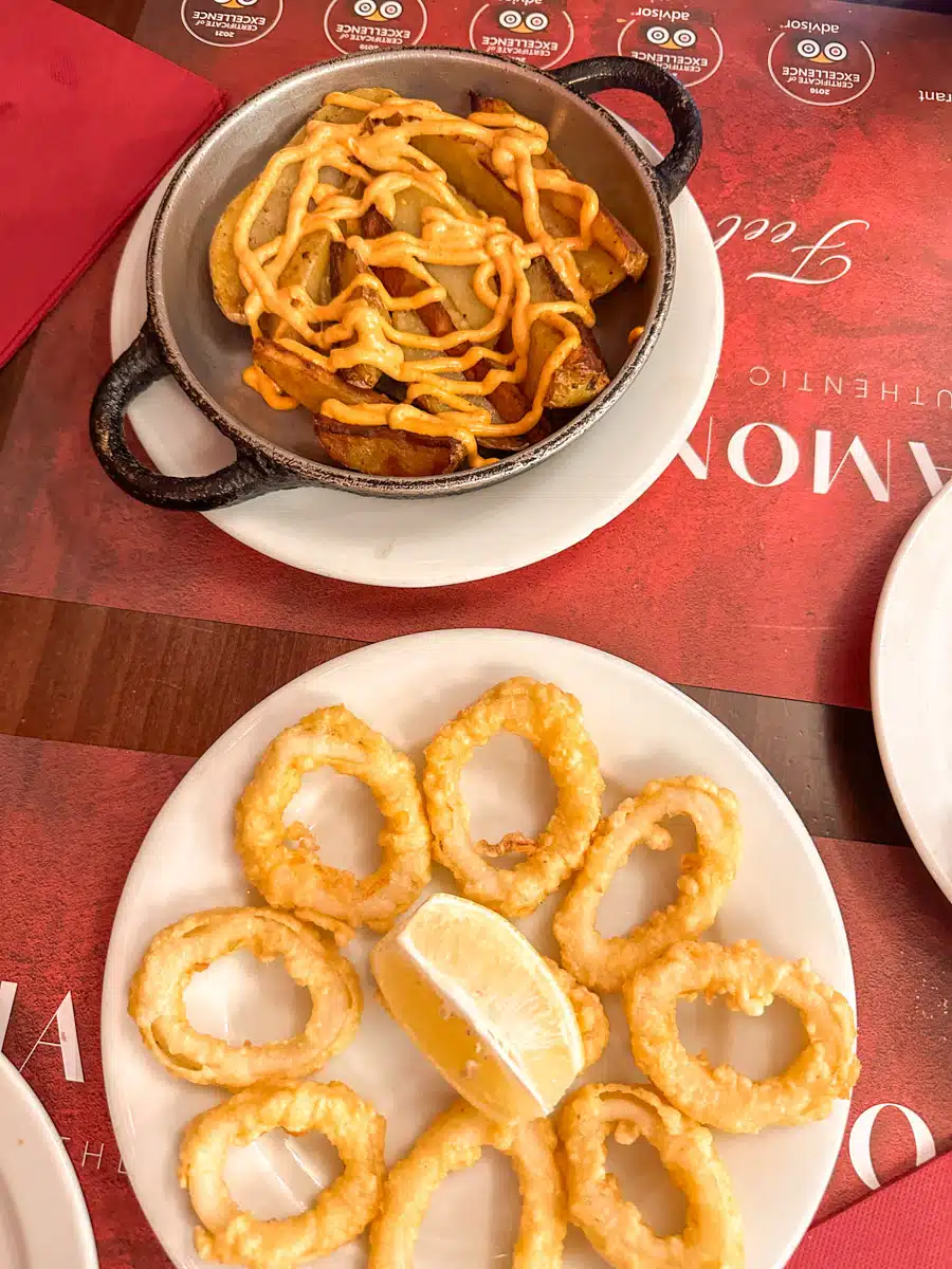 A table with traditional Spanish cuisine, featuring patatas bravas and crispy calamari rings, served with lemon and a spicy aioli.
