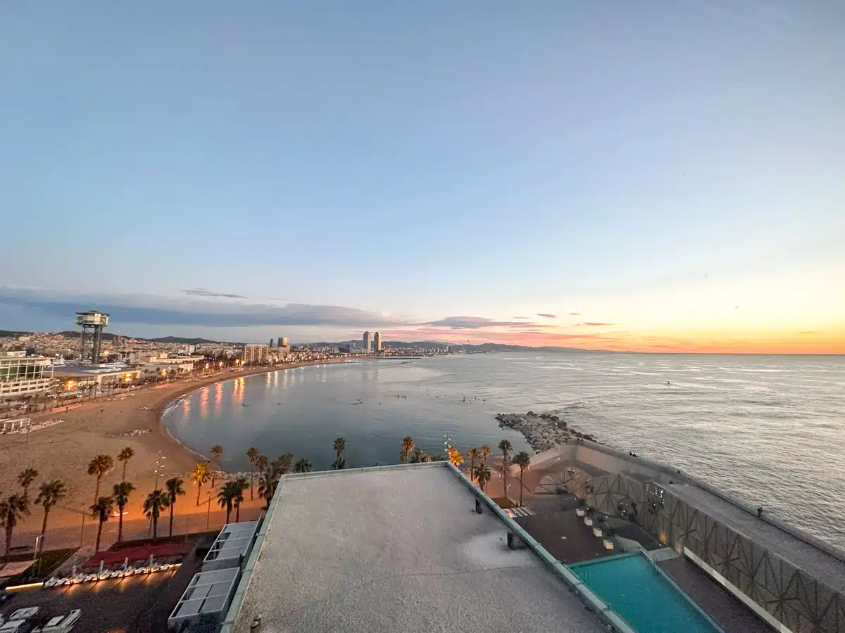 An elevated evening view from a hotel balcony showing a vibrant sunset over Barcelona's beach and cityscape.